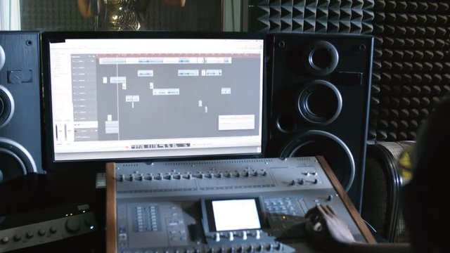 Tilt up of male audio engineer using mixing console and watching energetic young black woman in sparkly dress singing into microphone in isolation booth of recording studio