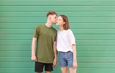 Beautiful young couple in a stylish casual dress standing on a green background, a boy kisses a girl on his cheek, she is satisfied.Young man kisses a girl on a turquoise background, love concept