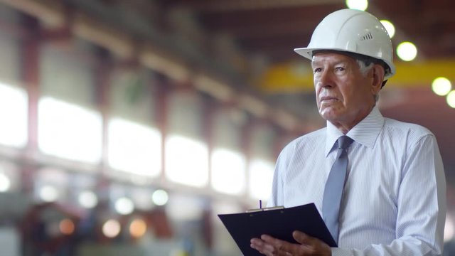 Chest-up shot of senior facility manager dressed in stripy shirt, tie and hardhat observing working process on factory floor, making notes on clipboard