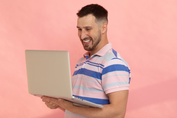 Studio closeup of smiling Caucasian man dressed in T-shirt, standing and holding laptop in his hands, browsing webpages, looking through photos and communicating online with pleasure.