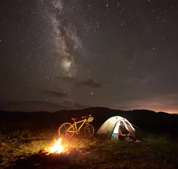 Gordijnen Young woman tourist having a rest at night camping near burning campfire, illuminated tourist tent, mountain bicycle under amazing beautiful evening sky full of stars and Milky way. Astrophotography © anatoliy_gleb