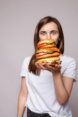 Studio portrait of young brunette woman in white t-shirt holding enormous burgers on her hand looking shocked or surprised at camera.