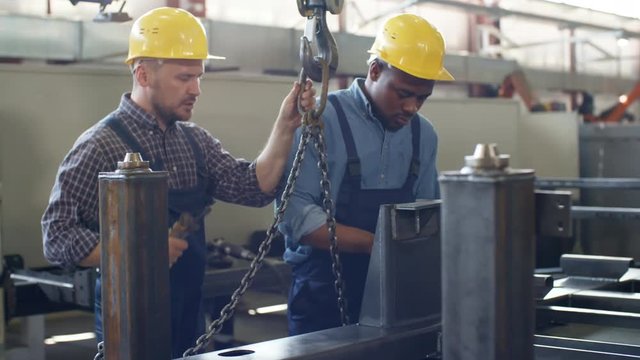 Panning medium shot of Caucasian crane operator instructing Afro-American trainee and fixing finished metal structure onto crane hooks and chains, both dressed in overalls and hardhats