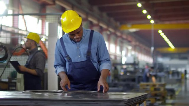 Thigh-up panning shot of Afro-American factory controller dressed in blue denim shirt, overalls and hardhat, inspecting batch of finished toothed gearwheels, with sprawling workshop in background