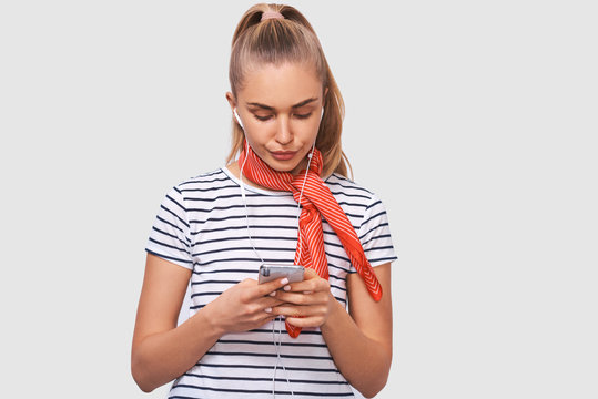 Displeased young woman using cell phone, messaging to her boyfriend. Unhappy female frowning her face and chatting with her boyfriend, looking at screen of smart phone, listening music in earphones.