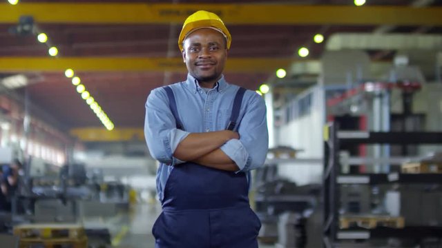Pan thigh up shot of black technician dressed in work overalls and safety helmet standing in busy sprawling workshop with folded arms