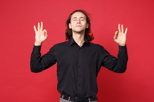 Stylish young curly long haired man in black shirt posing isolated on red wall background studio portrait. People sincere emotions lifestyle concept. Mock up copy space. Hold hands in yoga gesture,