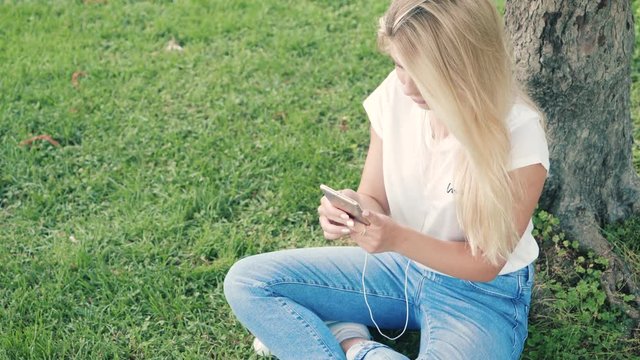 girl sitting in the park on the grass, listening music in earphones, chatting and shooting photo on smartphone. blonde young girl on summer day relaxing outdoors