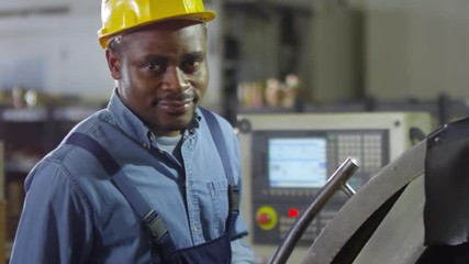 Close-up panning shot of black factory mechanic dressed in blue overalls and hardhat operating modern cutting lathe and smiling happily, with electronic control panel in background