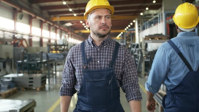 Dollying waist-up shot of Caucasian engineer strolling along factory floor, glancing around with serious, concerned face and frowning, with rows of metal components and machinery in background