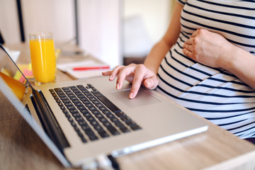 Close up of pregnant freelancer using laptop in home office. On desk laptop, orange juice and notebook. Working pregnant women concept.