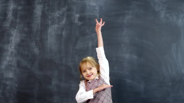 Portrait shot of adorable little schoolgirl standing against blackboard and smiling while raising her hand to answer question