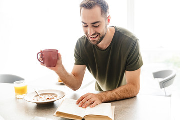 Fototapeta na wymiar Cheerful young bearded man indoors at the kitchen have a breakfast eat corn flakes with milk reading book drinking coffee.
