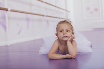 Fototapeta premium Little cute ballerina girl lying on the floor at ballet school, looking tired, copy space. Adorable little ballerina in tutu skirt, looking sad and tired, lying on the floor after exercising at ballet