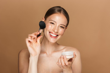 Happy beautiful young amazing woman posing isolated over brown chocolate background wall holding powder and makeup brush.