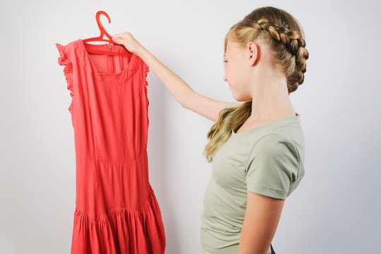 Teen girl choosing a dress for a fitting standing over grey background. Caucasian girl holding a red dress on a hanger.