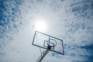 Basketball basket on a background of blue sky with light clouds. The sun is right above the basket like a ball. Place for text. The concept of summer sports, summer hobby.