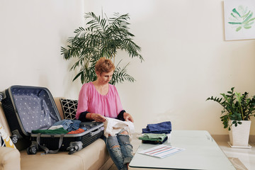 Senior woman sitting at table with plane ticket and stack of clothes and packing suitcase for summer vacation