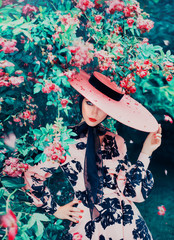 Fototapeta na wymiar charming girl hides part of her face under wide-brimmed pink hat with ribbons. Lady with warm make-up, red lips in garden of blooming roses , Girl flirts on happy valentines day