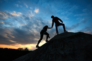 Silhouettes of two climbers reaching summit, one holding hand of partner assisting to make last step to top. Young man and woman athletes bouldering on high rock. Amazing sky and sunset on background.