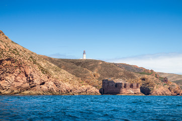 Coast of the Berlenga island, seen from the sea, with the Fort São João Baptista das Berlengas highlighted and in the background the lighthouse with the blue sky, on a summer day.