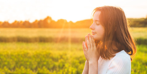 Teenager Girl closed her eyes, praying in a field during beautiful sunset. Hands folded in prayer...