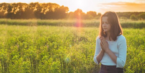 Naklejka na ściany i meble Teenager Girl closed her eyes, praying in a field during beautiful sunset. Hands folded in prayer concept for faith, spirituality and religion. Peace, hope, dreams concept