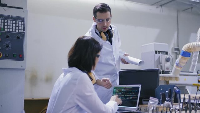 Two cnc engineers, male and female wearing white coats, protective eyewear and ear muffs around necks discussing operations on laptop computer screen in laboratory, tracking right shot