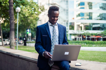 African American businessman holding laptop notebook wearing blue suit and using modern smartphone near office. Business concept