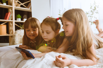 Girls using different gadgets at home. Childs with smartphone lying and smiling, watching videos or playing games. Making selfie, chating, gaming. Interaction of kids and modern technologies.