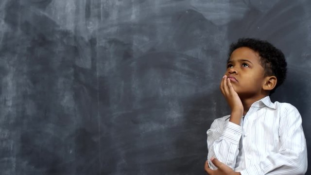 Portrait shot of cute little African-American boy standing before blackboard and thinking about something