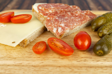 A slice of bread with salami and a slice of bread with cheese, while still small cocktail tomatoes and pickled cucumbers, served on a wooden board
