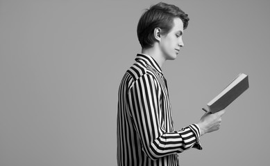 Young handsome man in striped shirt reading a book on gray background