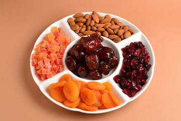 Mix of dried fruits and nuts on a white plate. Apricot, almond, raisin, dates fruit. On a brown background. Space for text or design.