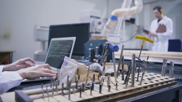 Closeup hands of female engineer in white coat working on laptop computer surrounded by tools in cnc lab, male technician checking laser engraving machine in background, tracking left side view shot