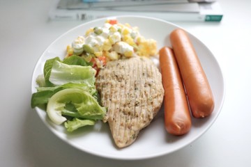 In selective focus a white dish of pepper chicken steak with fresh vegetables and corn carrot tuna salad on white table and blur a pile of magazines