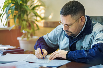 A male engineer in glasses in the office at the table signs the documents. Inspection, recording of prescriptions and violations in the log.