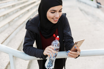 Young smiling muslim sports fitness woman dressed in hijab and dark clothes outdoors at the street with steps on background drinking water holding bottle.