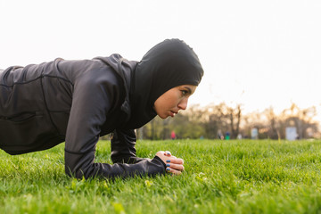 Young muslim sports fitness woman dressed in hijab and dark clothes outdoors in green nature park make exercises plank.