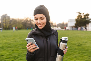 Young cheerful muslim sports fitness woman dressed in hijab and dark clothes outdoors in green nature park holding bottle with water using mobile phone chatting.