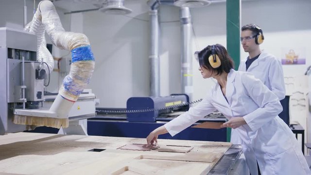 Two cnc engineers, male and female, in white coats, safety eyewear and ear muffs controlling laser engraving machine cutting wood in factory laboratory and checking ready elements, tracking right shot