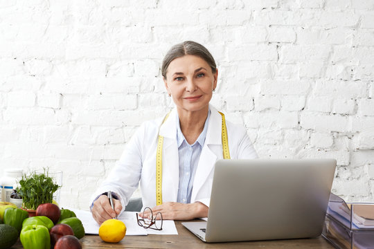 Picture of elderly mature female expert on diet and nutrition sitting at desk wearing white uniform and measure tape around her neck, using laptop, writing, making dieting plan for her client