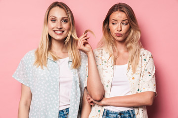 Sad woman holding hair of her happy pretty blonde women friend sister posing isolated over pink...