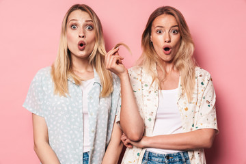 Surprised woman holding hair of her excited pretty blonde women friend sister posing isolated over pink wall background.