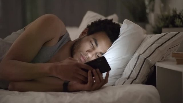 Medium shot of young black man lying in bed late at night and using smartphone application