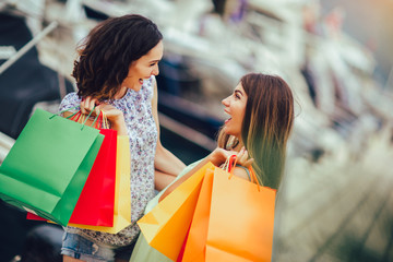 Female friends with shopping bags walking by the harbor of a touristic sea resort with boats on background