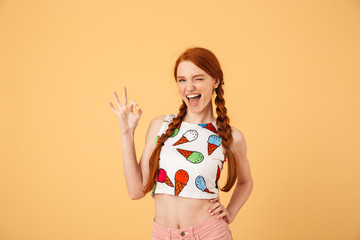 Young cheerful beautiful redhead woman dressed in ice cream printed t-shirt posing isolated over yellow background showing okay gesture.