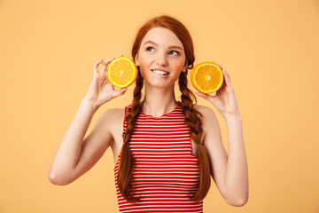 Happy young beautiful redhead woman posing isolated over yellow background holding orange.