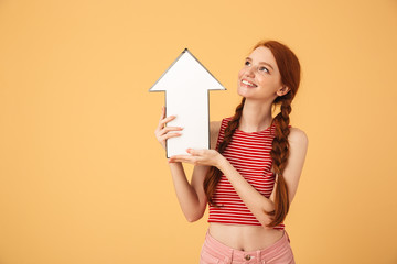 Cheerful  young beautiful redhead woman posing isolated over yellow background holding arrow.