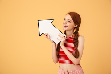 Happy  young beautiful redhead woman posing isolated over yellow background holding arrow.